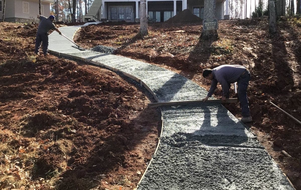 Two crew members screeding a curved concrete walkway on a graded slope up to a home